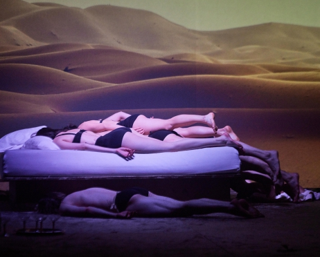 Na zdjęciu łóżko na którym leży kilka bezwładnych ciał kobiet i mężczyzn, Każdy z nich ma na sobie czarną bieliznę. W tle wyświetlane jest zdjęcie przedstawiające piaszczystą pustynię. Zdjęcie jest mroczne.